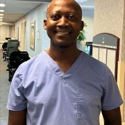 Portrait of Asare Bonsu, CNA at Holden Rehabilitation & Skilled Nursing Center, smiling in gray scrubs. Honored as September 2025 Employee of the Month.